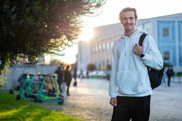  Happy man standing on a sidewalk near his faculty building with a backpack. Student smiling while...