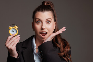 Surprised businesswoman holding a small yellow alarm clock, expressing shock at the time. Concept of time management, deadlines, and urgency
