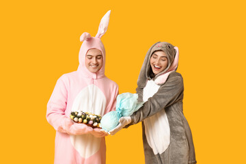Beautiful young couple in bunny costume with Easter gift egg and wreath on yellow background