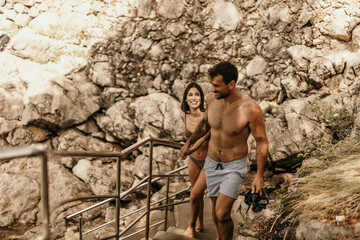Young, beautiful shirtless couple strolling towards a rocky beach, ready for a summer adventure