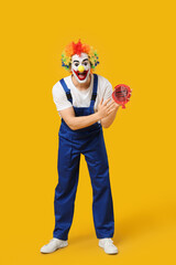 Screaming young man as clown in jumpsuit with whoopee cushion on yellow background