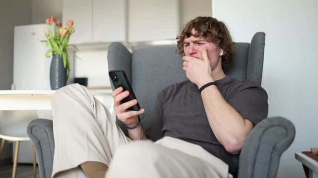 A young man sits in a chair, looking at his phone with a displeased expression as he covers his mouth, likely feeling nauseous or reacting to something unpleasant.