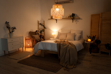 Interior of bedroom with beige plaid on bed, chest of drawers and glowing lamps at night