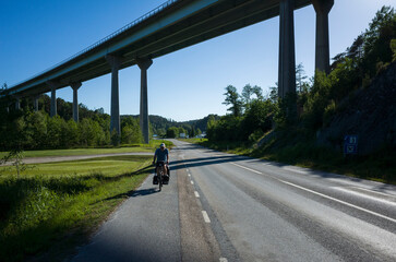 A man travels by bicycle through Bohusl&auml;n, Sweden, riding under the E6 highway bridge on a sunny day. A scenic road surrounded by green nature, symbolizing adventure and freedom
