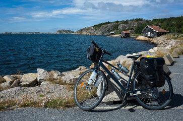 Obraz premium A touring bicycle with panniers leans against a rock at Kungsvik Harbor on Sweden’s west coast. A perfect summer stop on a cycling trip, with rocky shores, old boathouses, and blue water