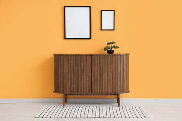 Commode with bonsai tree and blank frames on yellow wall in room