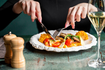 A close-up view of a woman dining elegantly, using a fork and knife to enjoy a plate of colorful vegetables and corn, with a glass of white wine nearby.