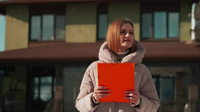 Professional realtor presenting new property, holding orange folder while standing confidently outdoors, wearing beige down jacket in suburban neighborhood during winter season