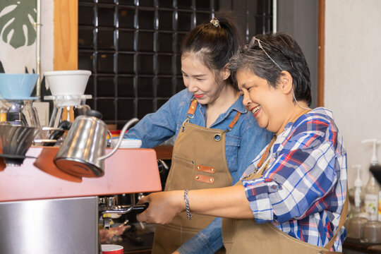 Asian mother and daughter bonding relationship small business lifestyle open cafe coffee shop after retirement, happy 60s elderly senior female laughing with young barista waitress learning technology