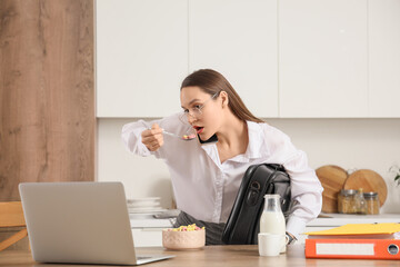 Hurrying young businesswoman talking by mobile phone while having breakfast in kitchen