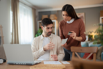 Obraz premium Young couple standing at home discussing documents while working on a laptop together. Concept of financial planning, teamwork, decision making and modern lifestyle indoors.