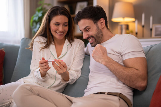 Cheerful couple looking at positive pregnancy test, sitting on bed