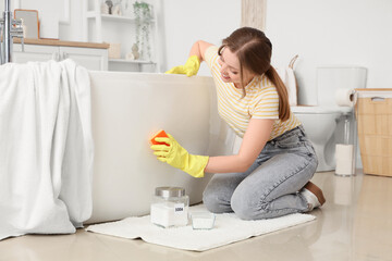 Young woman with sponge and baking soda cleaning bathtub in room