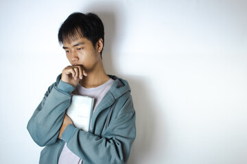 Asian teenager in a white t-shirt and blue hoodie, posing in deep thought while embracing a book