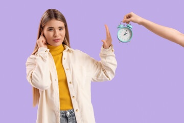 Displeased young woman rejecting hand with ringing alarm clock on lilac background