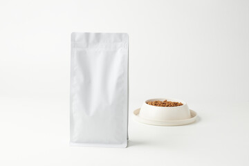White Packaging of Pet Food Stands Beside a Bowl of Kibble on a Neutral Background