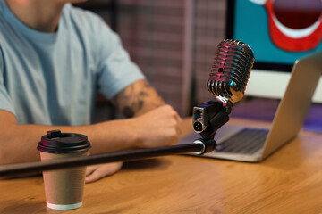 Microphone with coffee cup on table against man recording podcast in studio, closeup