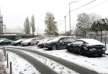 A serene snow-covered city street with trees draped in snow, parked cars lining the road, and faint...