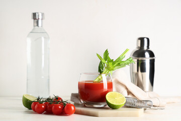 Glass of bloody mary with celery, cut lime, tomatoes and strainer on light background