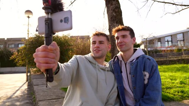 Young caucasian gay couple recording video with smartphone and external microphone, sitting on bench in a park