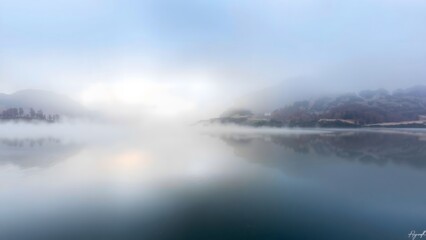A serene lake enveloped in fog reflects the surrounding mountains, creating a tranquil and ethereal atmosphere in soft, blue tones.  