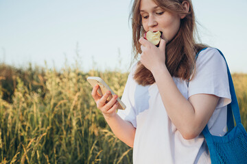 Fototapeta premium The girl is eating an apple and looking at the phone screen outdoors.