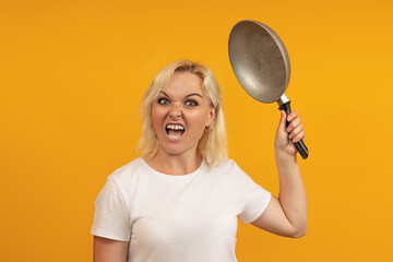 angry housewife with a frenzied face swings a frying pan towards the camera on a yellow background