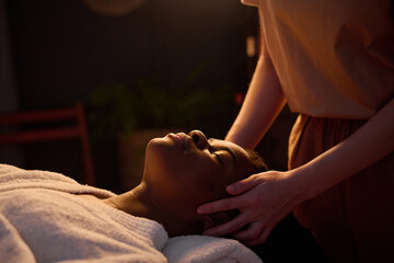 Medium close up of African American woman in bathrobe lying with closed eyes enjoying head massage...