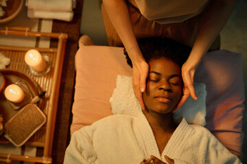 Top down shot of young African American woman in bathrobe having facial massage while lying on massage table