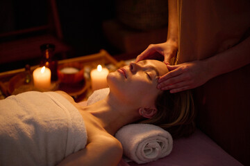 High angle of blonde woman lying on towel roll and getting facial massage by professional masseuse, room filling with aroma scent slowly