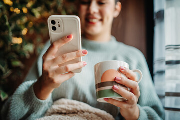 Young caucasian woman use phone send sms text messages and drink coffee by the window