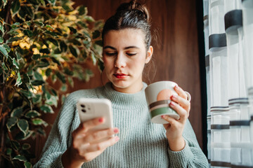 Young caucasian woman use phone send sms text messages and drink coffee by the window