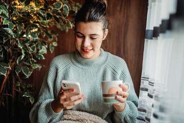 Young caucasian woman use phone send sms text messages and drink coffee by the window