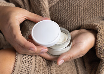 Closeup of woman in brown bathrobe opening white cream jar with hands, cosmetic mockup