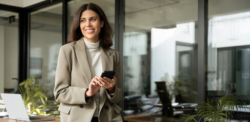 Smiling latin businesswoman it specialist working in modern office, holding smartphone cellphone. Middle eastern business woman in professional using mobile phone ai app for work . Banner, copy space