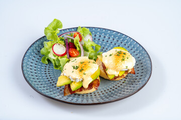 Delicious eggs benedict with avocado and bacon served on blue plate, accompanied by fresh salad of lettuce, cherry tomatoes, and radishes, creating vibrant and appetizing meal