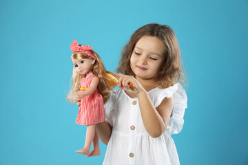 Cute little girl brushing doll's hair on light blue background