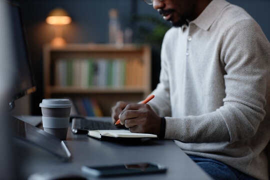 Cropped shot of Black man taking notes on small paper reminder managing working tasks and organizing time at desk in office, copy space