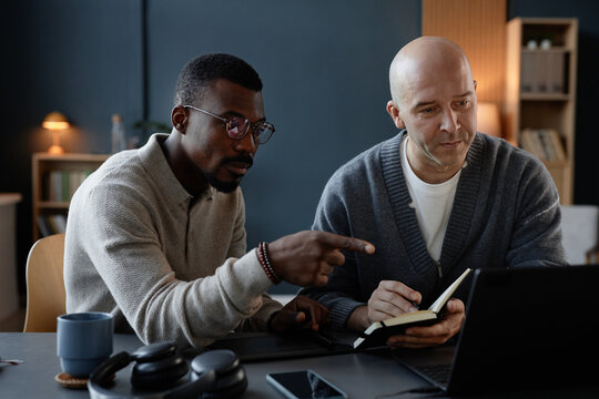 Black male interior designer pointing at laptop screen guiding trainee while male colleague writing in notebook, working at desk with digital devices for graphic arts in office