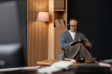 Wide angle shot of concentrated middle aged man with shaved head listening to music through headphones, while reading interior design magazine sitting in cozy home office