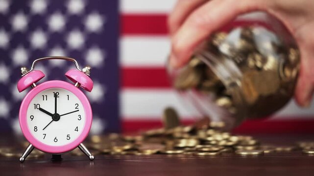A hand scatters gold money coins from a jar behind an alarm clock. USA flag in the background. Retirement, pension plannings, savings.