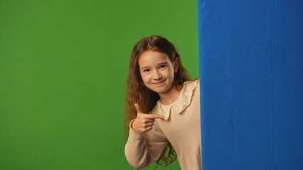 Little girl child peeks out and pointing at blue space, positive expression, isolated on chroma key green screen background mockup.