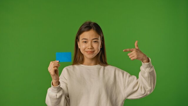 Asian girl child holding advertisement card and pointing finger at blue space, positive expression, isolated on chroma key green screen background mockup.