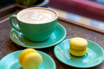 Fresh delicious coffee latte with pattern and two yellow macaroons in green dish on table. Selective focus