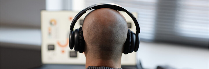 Website header shot of bald man shot from rear view listening to podcast through headphones while...