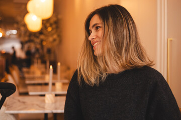 Happy young woman sitting on chair at cafe and looking at side. Portrait of comfortable woman in winter clothes relaxing on armchair. Portrait of beautiful girl smiling and relaxing.