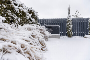 Snow-covered outdoor heat pump beside frosted bushes and conifers against a gray fence, illustrating winter home heating in a garden setting.