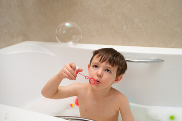 Baby boy blowing soap bubbles while taking a bath in a white bathtub. Playful child with wet hair enjoying bath time. Childhood fun and hygiene concept. A kid aged three years