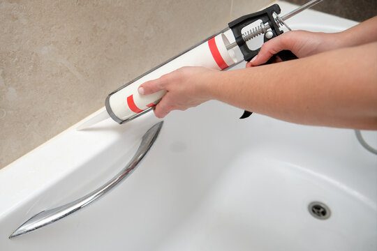 Hand applying sealant to bathtub corner using caulking gun. Home renovation and bathroom maintenance concept.