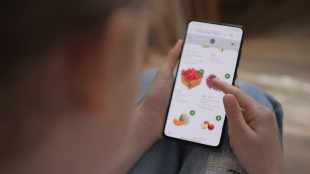 Woman is Shopping Online Food Delivery Market From Home. Girl Looks at Food in Grocery Online Store Sitting on Stairs at Home. Ordering Organic Food in Online Stores Using Mobile Phone Smartphone.